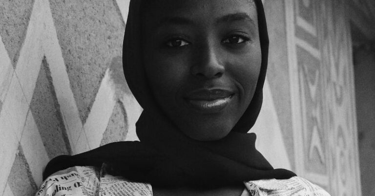 Documentaries - Black and white low angle of young smiling African American female in shawl on head and t shirt with text pattern standing leaned on concrete wall and looking at camera