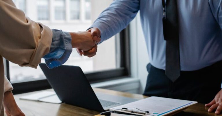 Collaborations - Crop unrecognizable coworkers in formal wear standing at table with laptop and documents while greeting each other before meeting